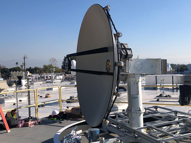 Megalodon dimensionally tunable reflector is deployed in the field. Hardware is on a rooftop, and there is a blue sky and mountains in the distance. The reflector is attached to a large column with electronics equipment and wiring visible. The image view is looking at the side of the reflector with interior of the dish and the edge of the deformation grid visible.
